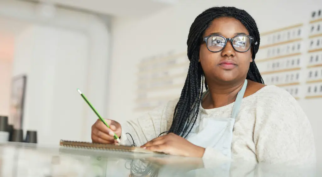 A young woman in glasses at a coffee shop looking out the window while writing something in a notepad.