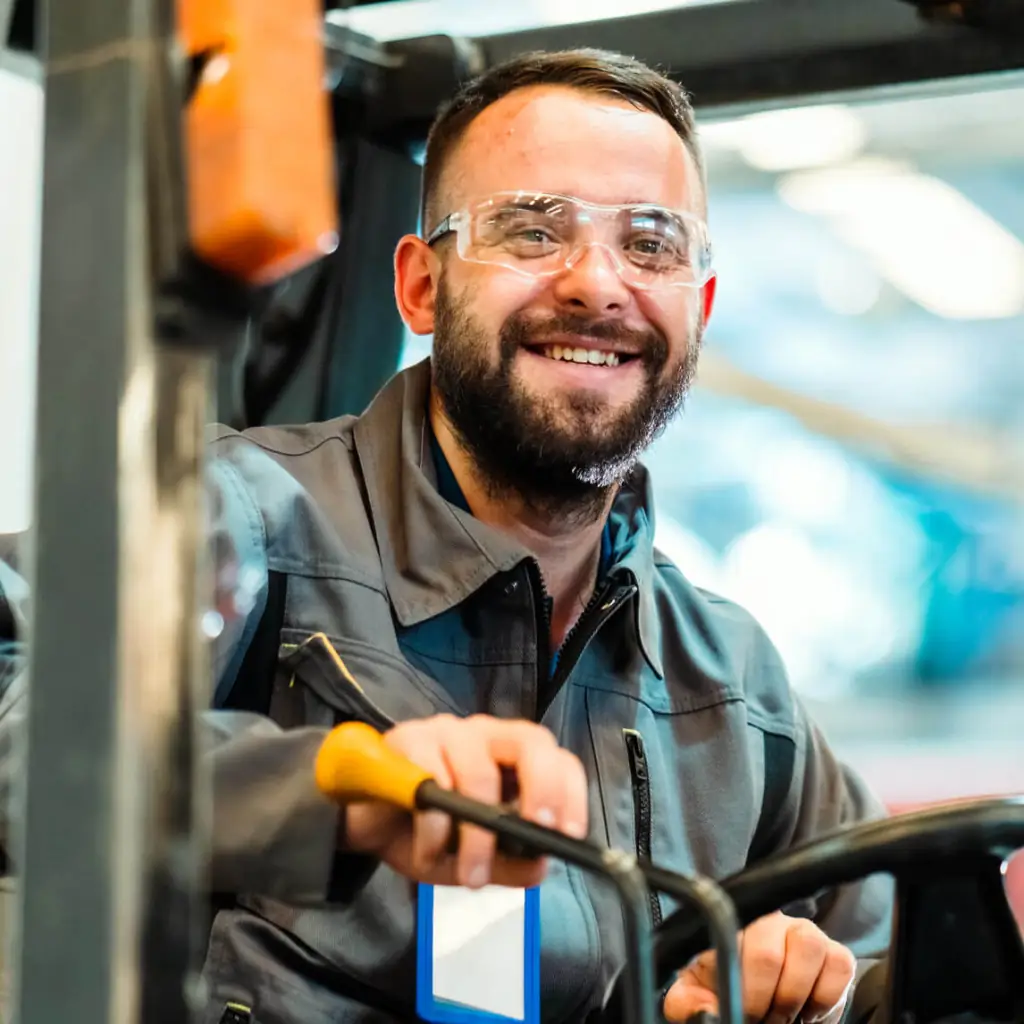 A smiling, bearded man wearing safety goggles operating a fork lift.