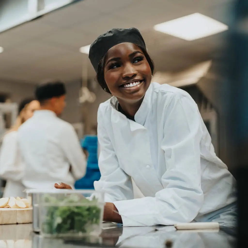 A young woman in a chef uniform leaning over a kitchen workstation that has food and a spatula on it.