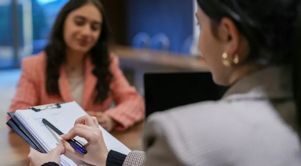 A woman pointing at lines on a notepad, explaining information to another young woman.